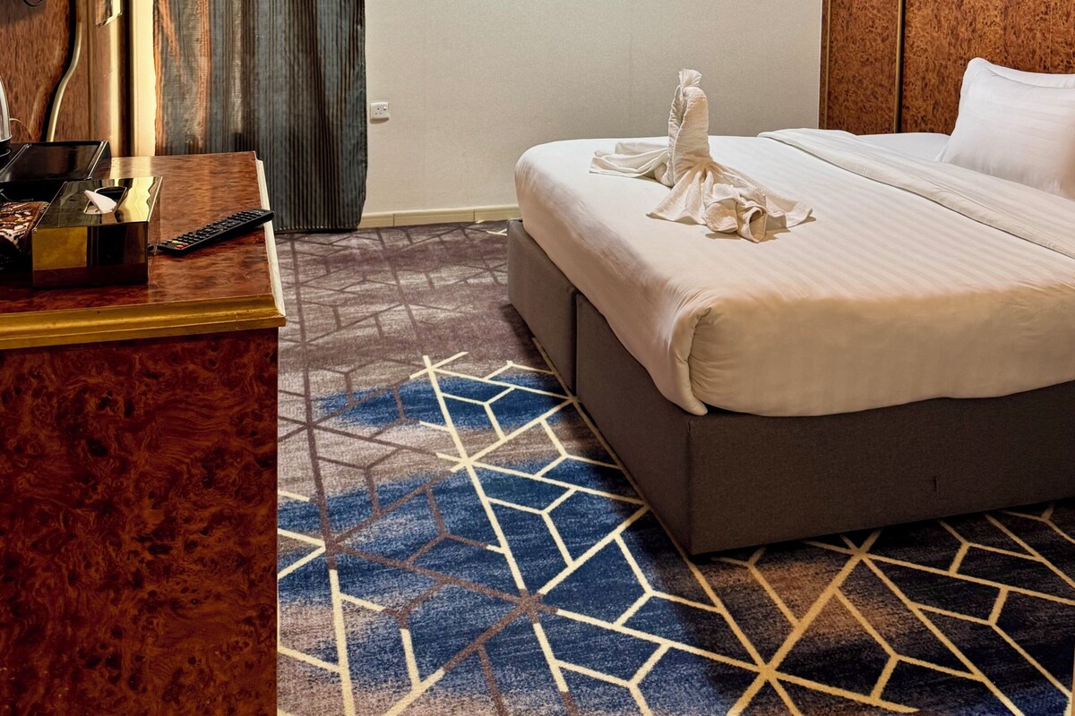 Habitación de hotel con una cama, un escritorio con televisor, control remoto y una alfombra estampada.
