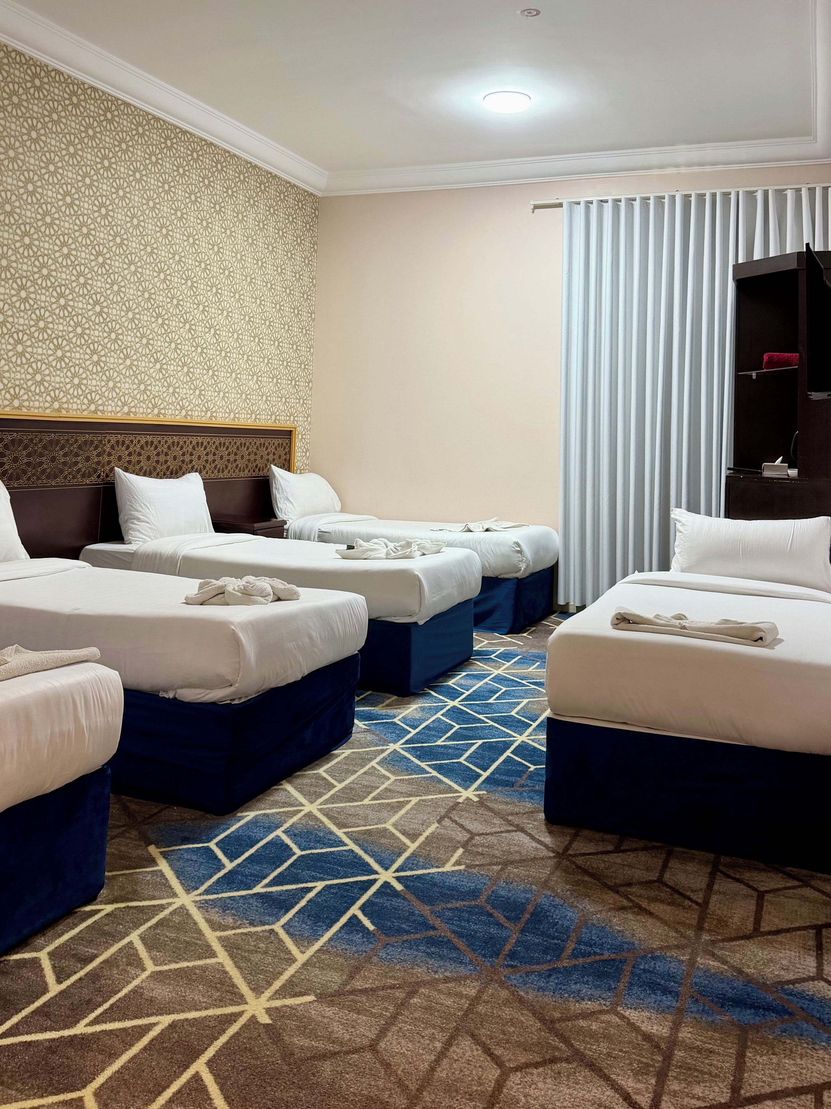 Una habitación de hotel con cuatro camas dispuestas en fila, alfombra estampada y un ventanal con cortinas.