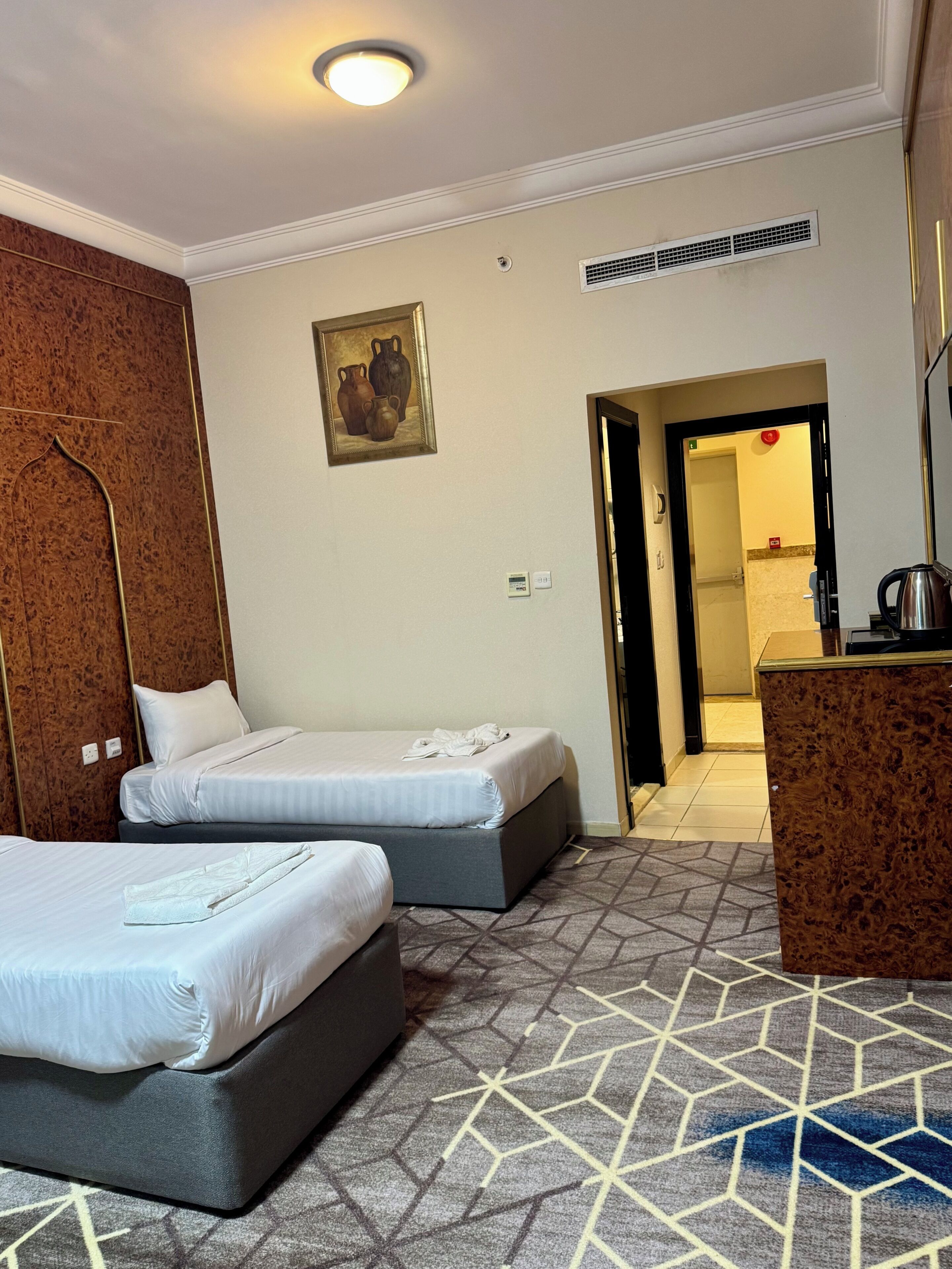 Habitación de hotel con dos camas, un armario de madera, un cuadro, un radiador y un baño visible a través de una puerta abierta.