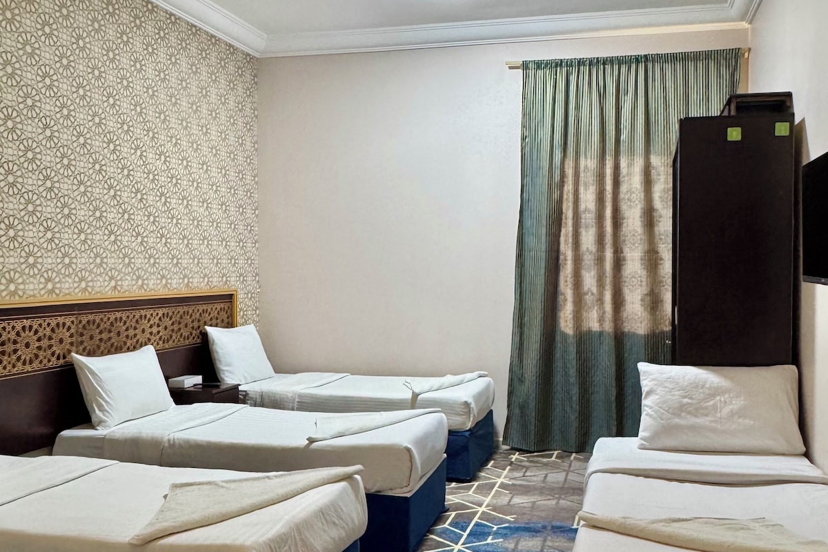 Una habitación de hotel con cuatro camas dispuestas en fila, un papel tapiz estampado, una lámpara en el techo, una cortina y un mueble negro.