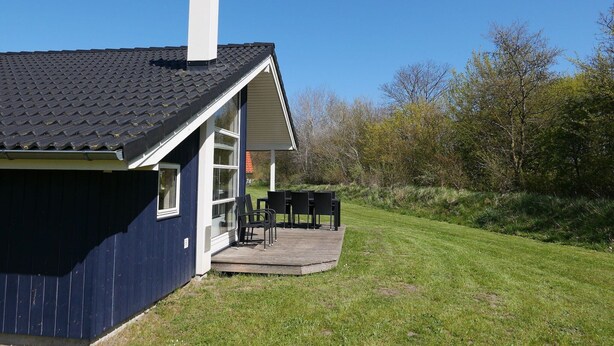 A house with a dark blue exterior, a white chimney, and a covered outdoor seating area with chairs and tables. - Ferienresort-Lodge in Großenbrode (Großenbrode)