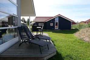 A deck with two lounge chairs and a table, overlooking a grassy area with a building in the background. - Familienversteck für 6 in Großenbrode (Großenbrode)
