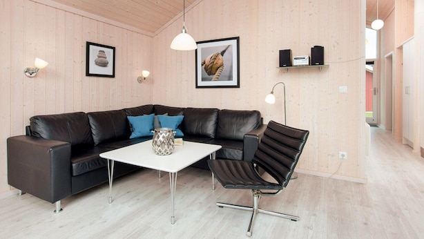 A modern living room with a leather sofa, a white coffee table, and a black chair. There are framed pictures on the wall and a shelf with speakers. - Chalet im skandinavischen Stil für 6 Personen (Großenbrode)