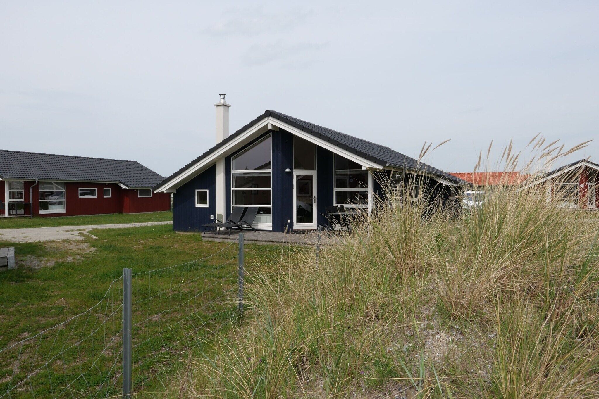 Een laag gebouw met een donkere gevel en een schuin dak, omringd door hoog gras en een hek.