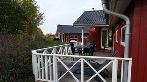Um deck de madeira com guarda-corrimão branco, uma mesa com cadeiras e um edifício vermelho ao fundo.