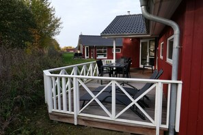 A wooden deck with a white railing, a table with chairs, and a red building in the background. - Geräumiges Strandhaus für 10 Personen (Großenbrode)