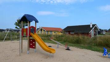 Ein Spielplatz mit Rutsche und Schaukeln auf sandigem Untergrund, umgeben von Häusern.