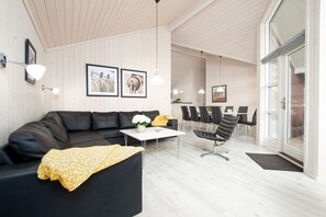 Ein modernes Wohnzimmer mit einem schwarzen Sofa, einem weißen Couchtisch und einer gelben Decke. An der Wand befinden sich gerahmte Bilder, ein Essbereich mit Stühlen und ein großes Fenster.