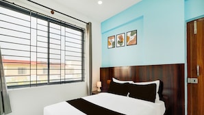 Quarto de hotel com uma cama grande, janela com persianas, cabeceira de madeira, luminária fixada na parede e porta com puxador.