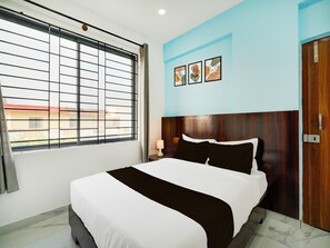 Ein Hotelzimmer mit einem großen Bett, einem Fenster mit Jalousien, einem Holzkopfschild, einer Wandlampe und einer Tür mit Griff.