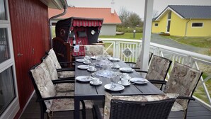 Ein Esstisch für sechs Personen auf einer Terrasse, mit Korbstühlen, ein rotes Gebäude im Hintergrund und ein gelbes Haus gegenüber der Straße.