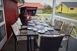 Ein Esstisch für sechs Personen auf einer Terrasse, mit Korbstühlen, ein rotes Gebäude im Hintergrund und ein gelbes Haus gegenüber der Straße.