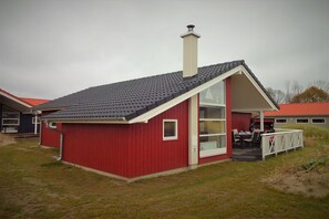 A red single-story building with a sloped roof, a chimney, and a large glass door leading to an outdoor seating area. - Serene Wellness Lodge für 8 Personen (Großenbrode)