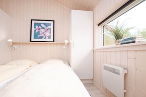 A bedroom with a bed, a framed picture on the wall, a radiator, and a window with a plant and books on the sill. - Großenbrode Meeresflucht (Großenbrode)