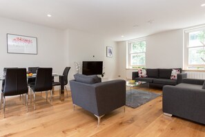 Een moderne woonkamer met een eethoek, grijze banken, een flatscreen televisie en grote ramen.