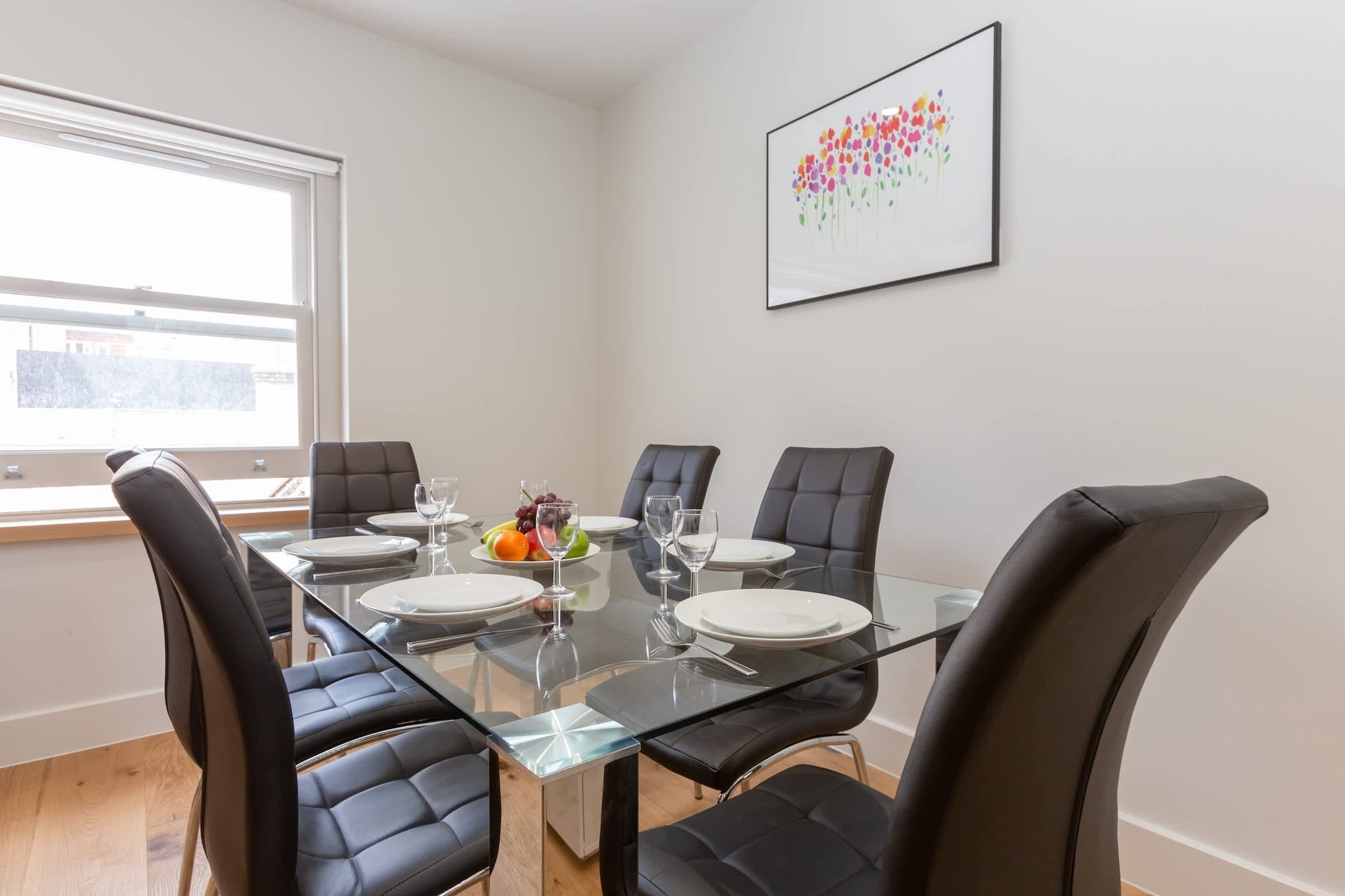 A dining area with a glass table and black leather chairs, a fruit plate, and a framed artwork on the wall.