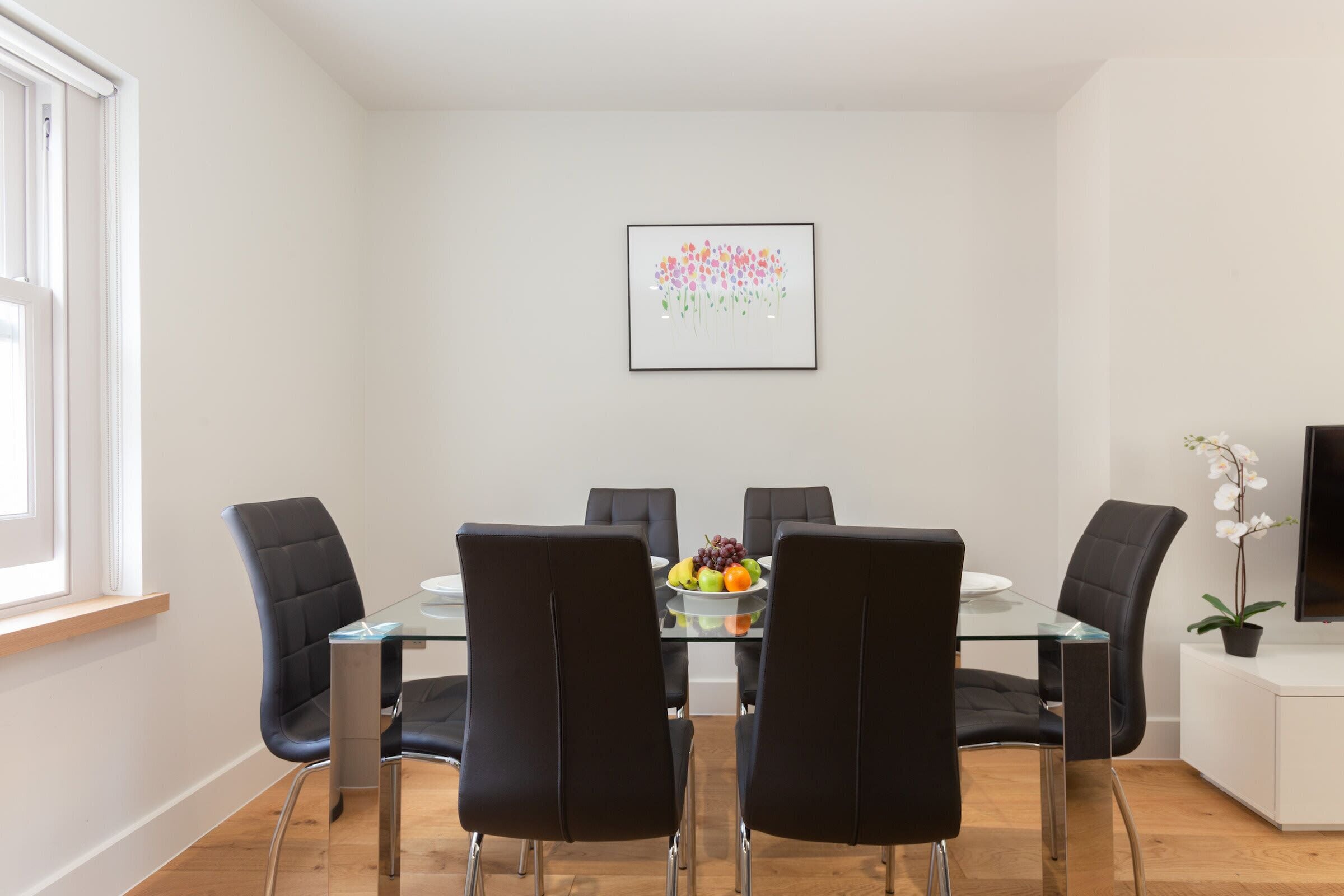 A modern dining area with a glass table, black chairs, and a fruit bowl.