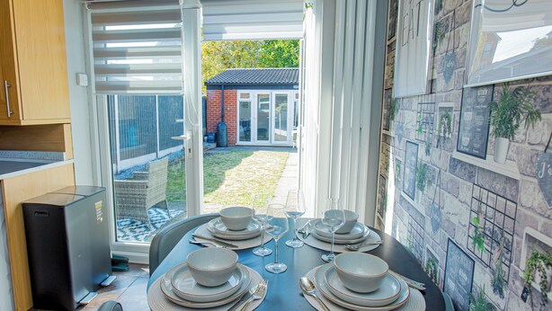 Comedor con una mesa dispuesta para cuatro, zona de cocina con lavavajillas y vista a un jardín a través de un ventanal.