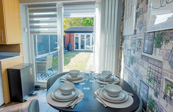 Un coin repas avec une table dressée pour quatre personnes, une cuisine équipée d’un lave-vaisselle et une vue sur un jardin, accessible par une porte vitrée.