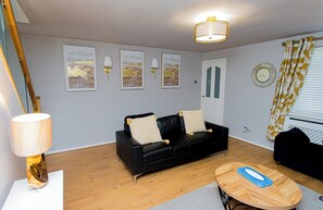 Ein Wohnzimmer mit einem schwarzen Sofa, zwei Wandlampen, einem Holztisch und einer Tür mit Fenster.