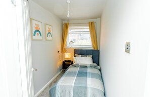 Ein Schlafzimmer mit einem Bett, einem Nachttisch mit Lampe, einem Fenster mit Vorhängen und zwei Wandbildern.