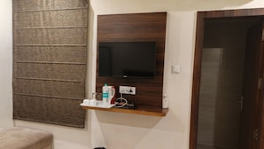 A room with a mounted TV, a small shelf with a bottle, glasses, and a kettle, a trash bin, and a door. - DREAM INN (Lucknow)