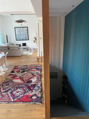 A living room with a colorful rug, a sofa, a dining area with chairs, and a staircase leading to another level. - The Place to Be: Monticelli Square (Marseille)