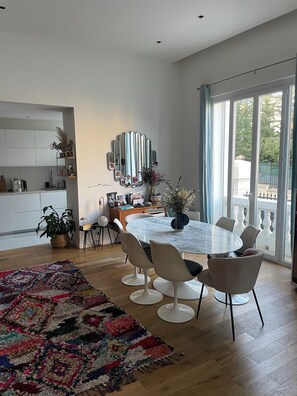 A modern dining area with a round table, white chairs, a colorful rug, and a large window with a view of greenery. - The Place to Be: Monticelli Square (Marseille)