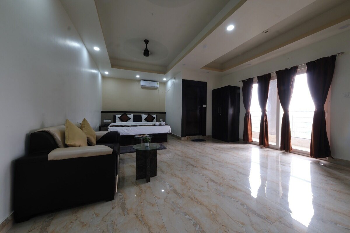 Habitación de hotel con cama, sofá, mesa de centro y una ventana grande con cortinas.