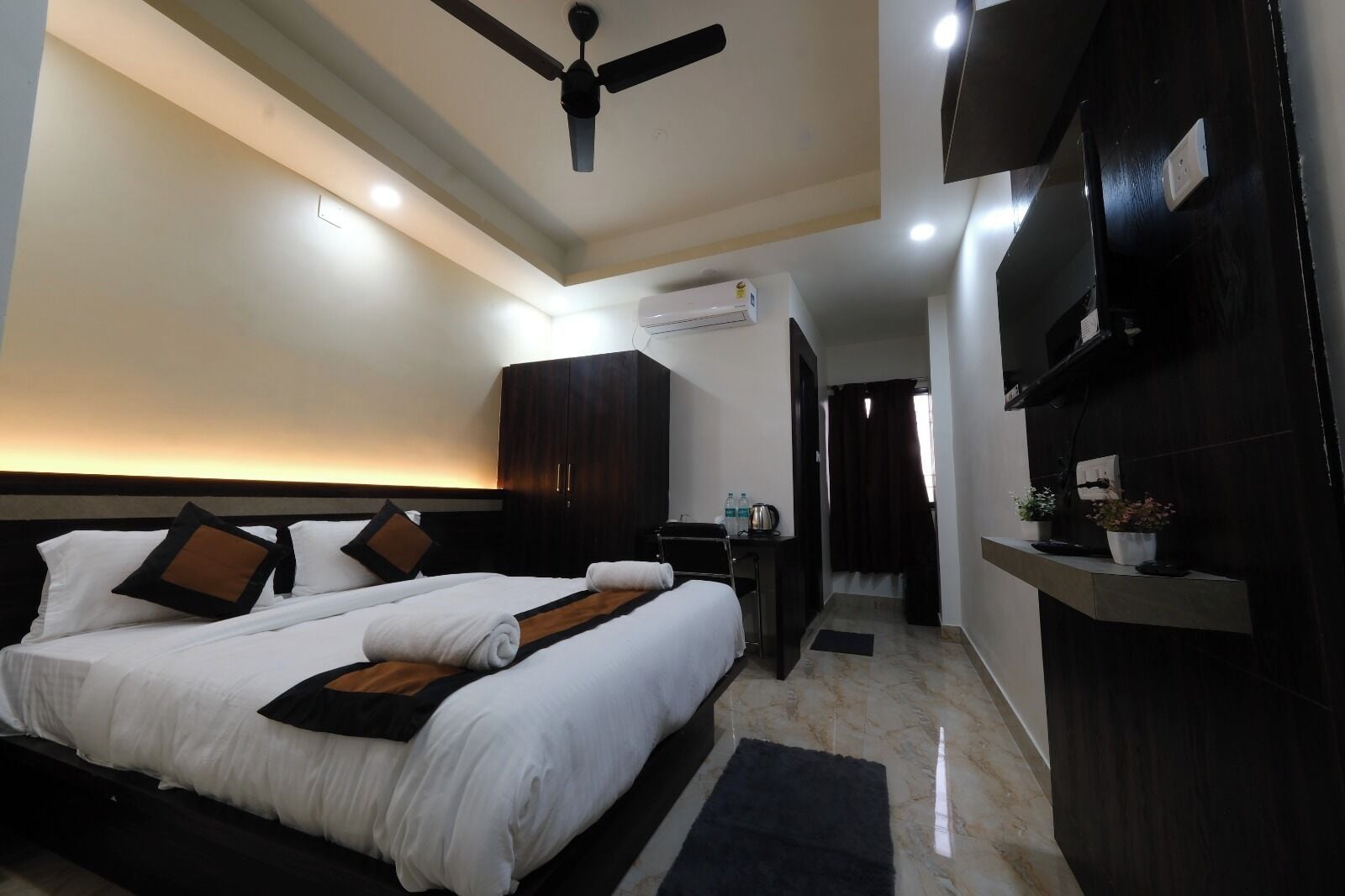 Ein Hotelzimmer mit Bett, Deckenventilator, Klimaanlage, Schreibtisch mit Stuhl und an der Wand montiertem Fernseher.