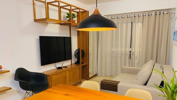 A modern living room with a wooden dining table, a black chair, a sofa, a TV mounted on the wall, and a ceiling fan. - Deluxe Apartment with Sea View & Pool Cumbuco (Praia de Cumbuco)