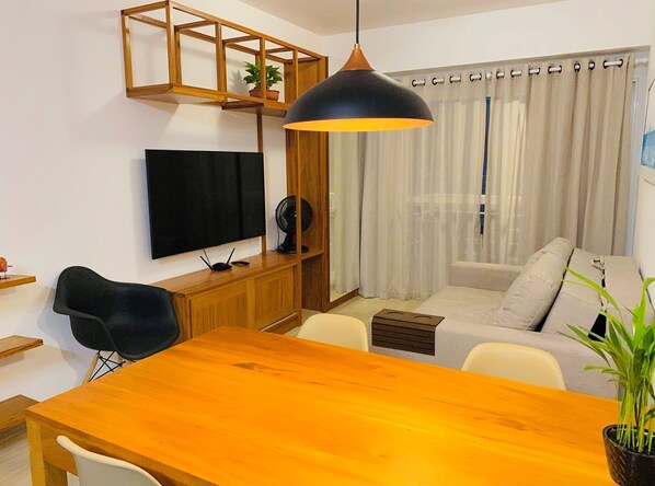 A modern living room with a wooden dining table, a black chair, a sofa, a TV mounted on the wall, and a ceiling fan.