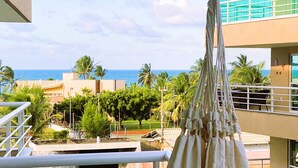 A balcony with a view of the sea, palm trees, and a building with a blue facade. - Deluxe Apartment with Sea View & Pool Cumbuco (Praia de Cumbuco)