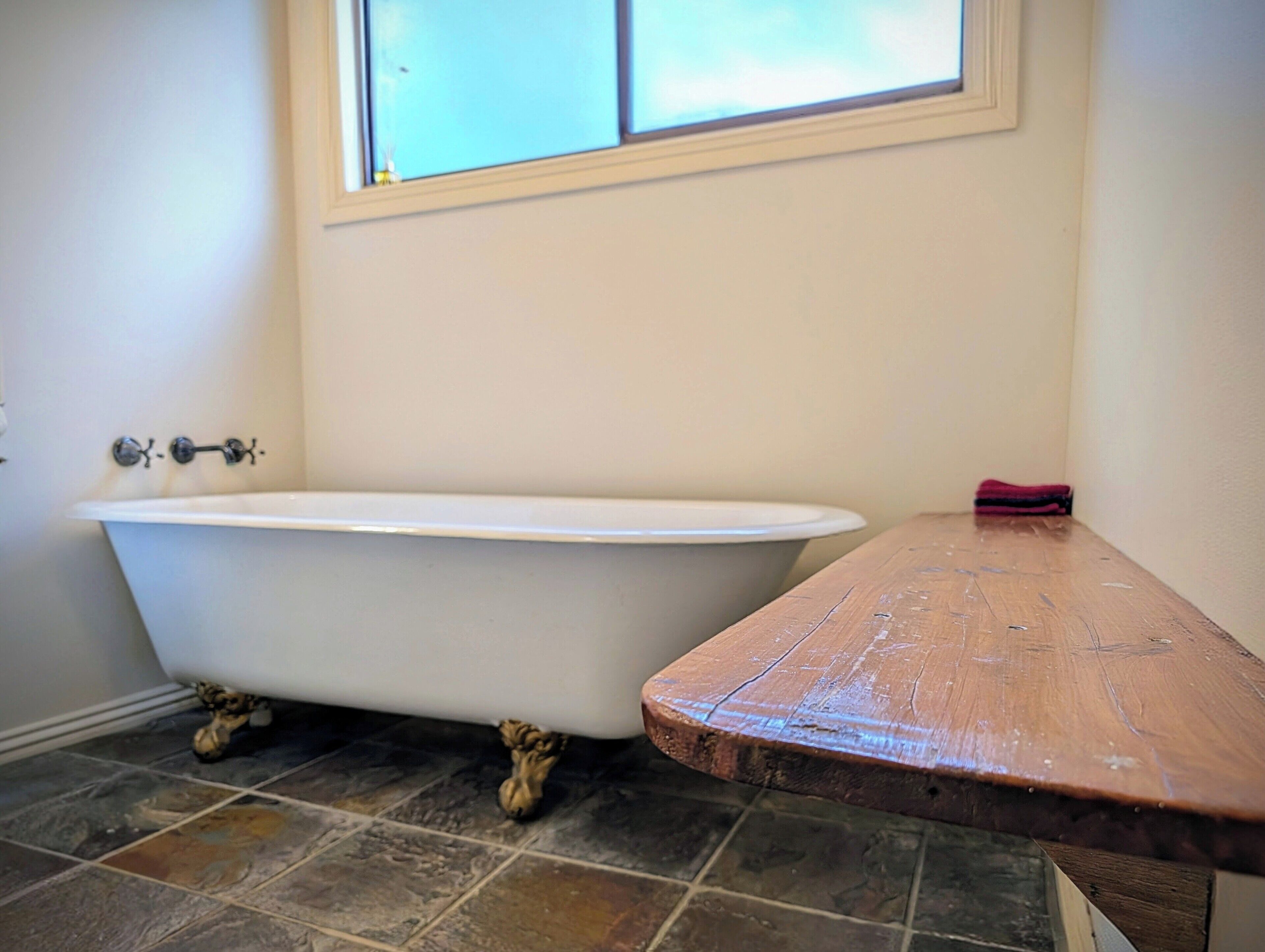 Une salle de bain avec une baignoire blanche, une fenêtre située juste au-dessus et un banc en bois.