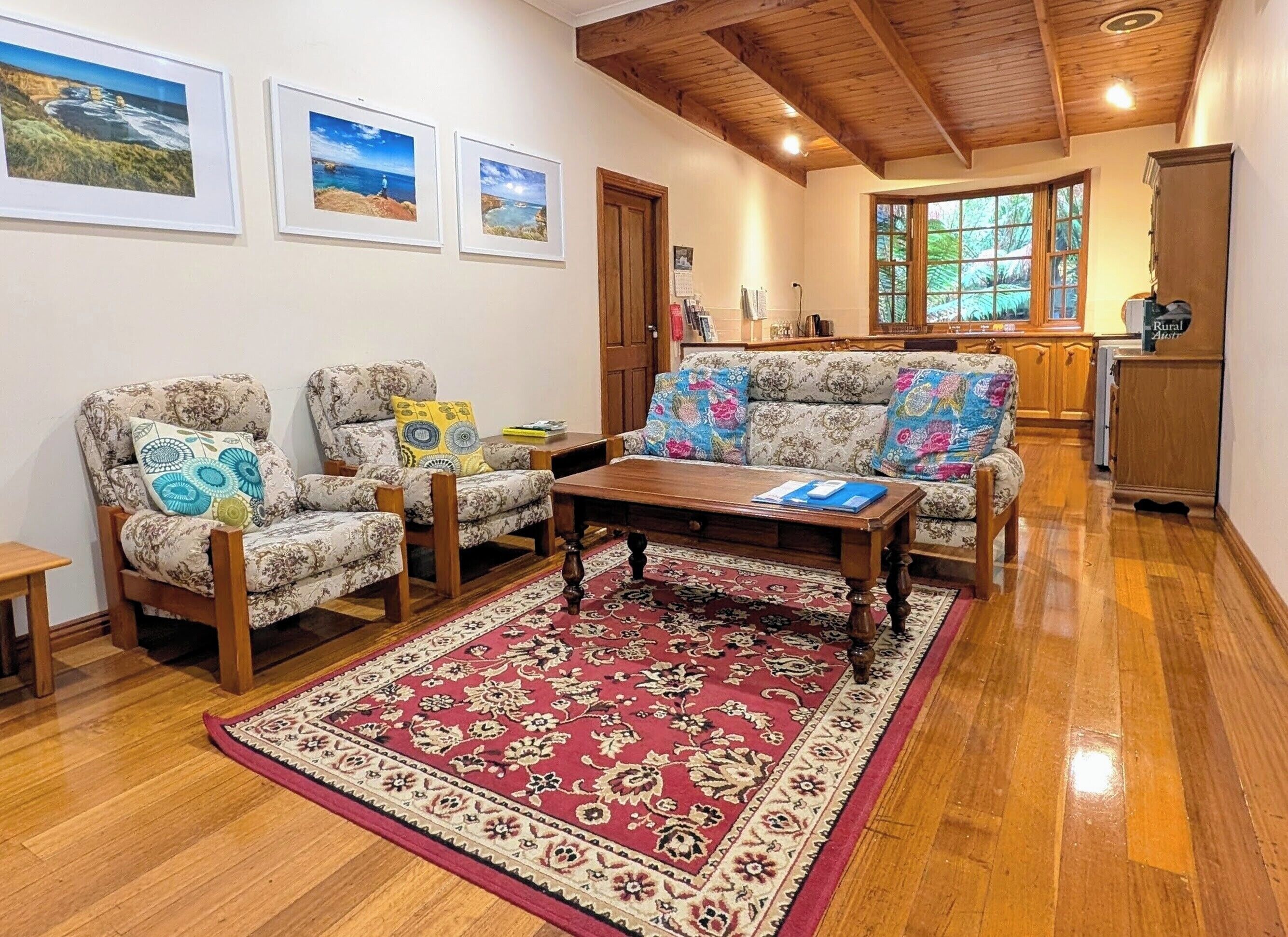 A living room with two patterned armchairs, a wooden coffee table, a red patterned rug, and framed coastal landscape pictures on the wall.