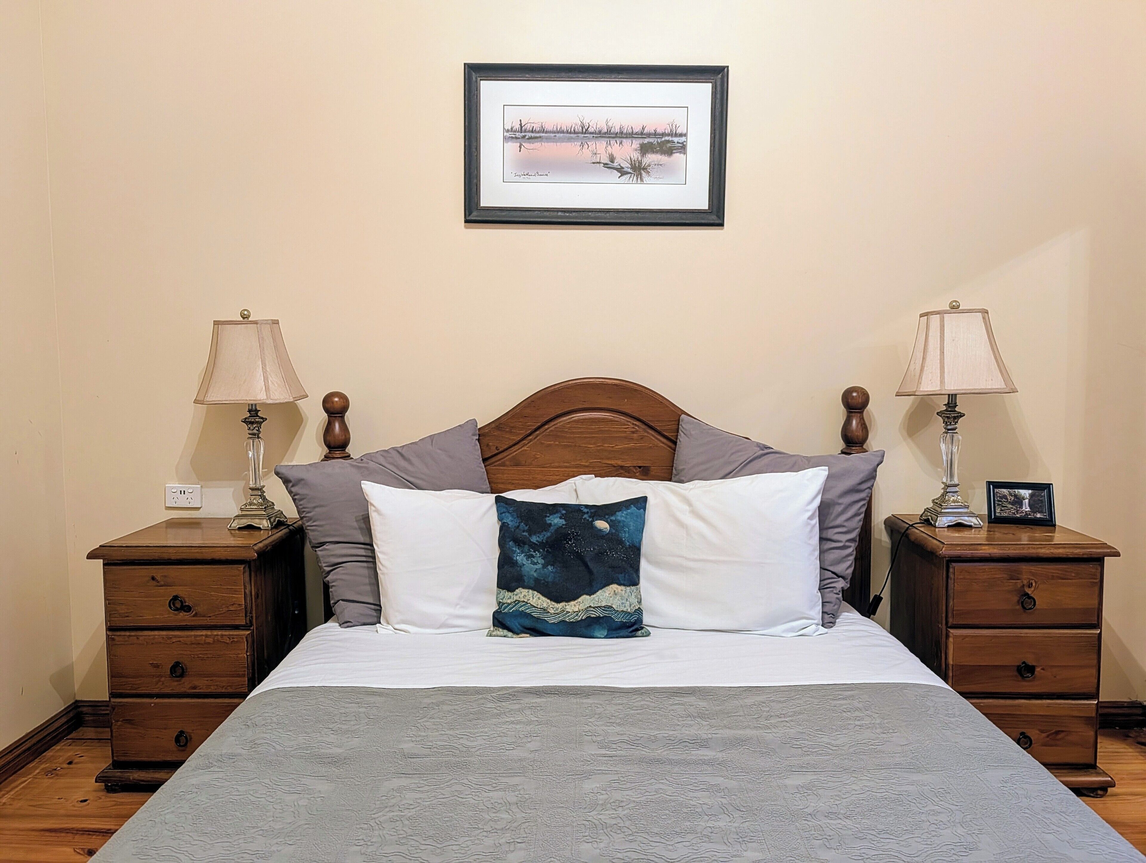 A bedroom with a wooden headboard, a bed with white and patterned bedding, two bedside tables with lamps, a framed picture on the wall, and a small drawer unit.