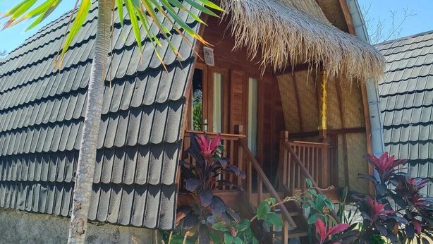 A thatched-roof bungalow with a wooden porch, surrounded by tropical plants. - Cozzy Bilva Garden (Klungkung)