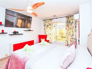 Ein Schlafzimmer mit einem Bett, einem an der Wand montierten Fernseher, einem Deckenventilator und einem Fenster mit Vorhängen.
