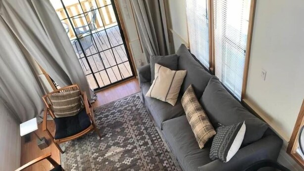 Una sala de estar con un sofá, un tapete estampado, una silla de madera y vistas a través de un ventanal.