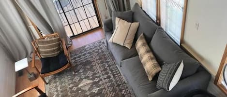 A living room with a sofa, a patterned rug, a wooden chair, and a view through a glass door.