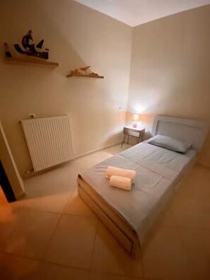 A bedroom with a bed, a bedside table with a lamp, a radiator, and a shelf with decorative items. - Quiet apartment in Artemis, Athens Airport (Artemis)