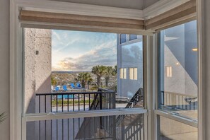 A balcony with a view of a pool and palm trees, a staircase, and a building with a textured facade. - 2BR Steps to Beach & Pool | Good Morning Rentals (Jacksonville Beach)