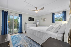 Ein Schlafzimmer mit einem Bett, einem Fernseher, einem Deckenventilator und Blick auf Palmen durch das Fenster.