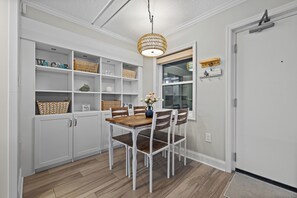 Een eetgedeelte met een houten tafel en stoelen, witte kasten met planken, en een hanglamp.