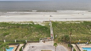 Vista a la playa con un camino que lleva a una zona de piscina, rodeada de vegetación y un cielo despejado.
