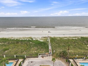 Blick auf einen Strand mit einem Weg zu einem Poolbereich, umgeben von viel Grün und blauem Himmel.
