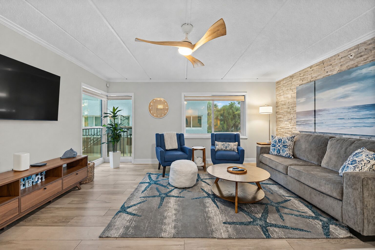 A modern living room with a ceiling fan, a large flat-screen TV, a grey sofa, two blue armchairs, a coffee table, and a large window with a view of palm trees.