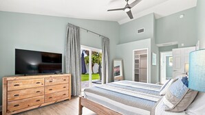 A bedroom with a bed, a TV on a wooden dresser, a ceiling fan, and a view of the outdoors. - Modern 3BR Near Beach | Good Morning Rentals (Jacksonville Beach)