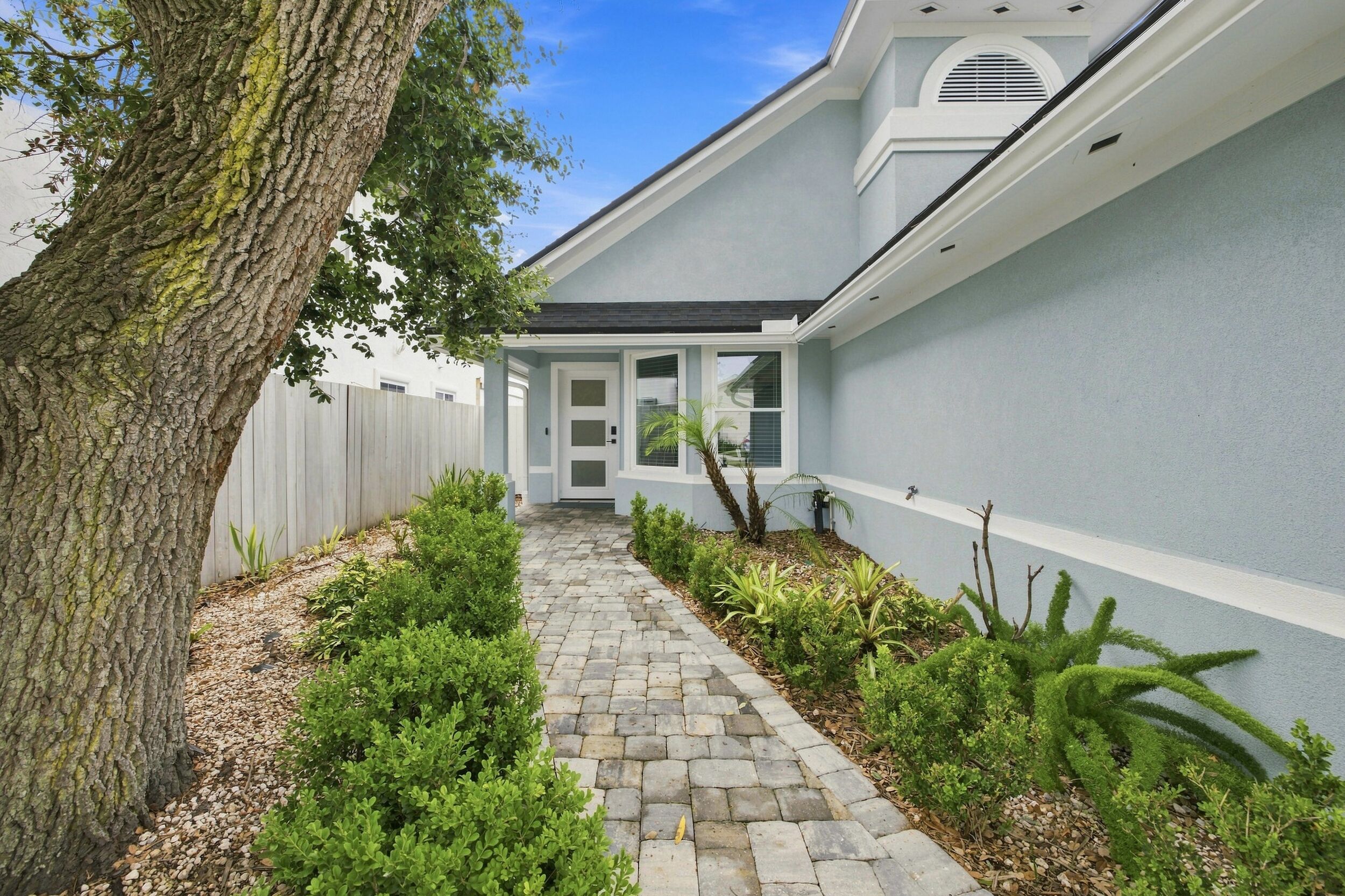 A pathway leading to a house with a white exterior, a wooden fence, and a tree on the left.
