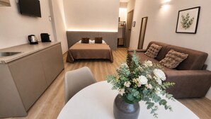 A compact hotel room with a kitchenette, a bed with a headboard, a sofa, and a dining table with a vase of flowers. - ROOM 1 Studio apartment near downtown and the train station, with private parking (TORINO)